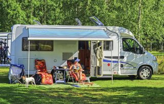Motorhome camping in campground summertime in Spain, woman sitting and relaxing on chair and reading book. Small dog is with her looking around. Mobile homes tent is opened and garden chairs and table is placed outside car. Campers sunroofs are opened because of warm weather.