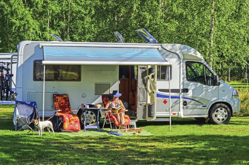 Motorhome camping in campground summertime in Spain, woman sitting and relaxing on chair and reading book. Small dog is with her looking around. Mobile homes tent is opened and garden chairs and table is placed outside car. Campers sunroofs are opened because of warm weather.