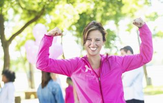 Mature adult Caucasian woman is smiling at camera and showing her muscles and strength. She is a breast cancer survivor or family member of survivor. She's wearing pink athletic clothing and a pink breast cancer awareness ribbon. She's a competitor in a breast cancer awareness marathon or 5k charity race.