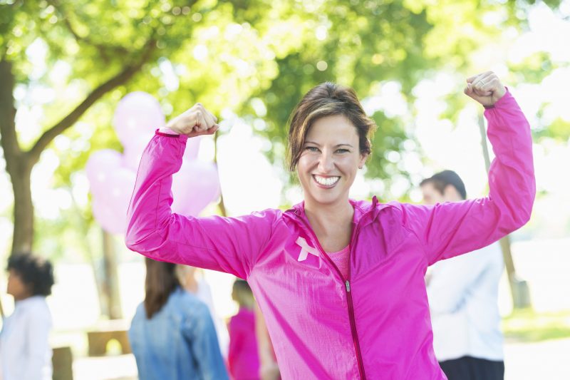 Mature adult Caucasian woman is smiling at camera and showing her muscles and strength. She is a breast cancer survivor or family member of survivor. She's wearing pink athletic clothing and a pink breast cancer awareness ribbon. She's a competitor in a breast cancer awareness marathon or 5k charity race.